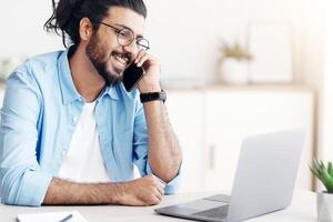 Remote Career. Smiling Eastern Freelancer Guy Talking On Cellphone And Working On Laptop At Home Office, Enjoying Distance Work Opportunities, Sitting At Desk, Looking At Computer Screen, Free Space photo