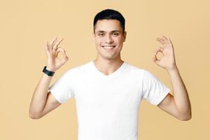 Great review, best offer, bloggers advice and advertisement. Attractive guy student in white t-shirt smiling and showing ok sign with hands, isolated on beige background, studio shot, free space photo