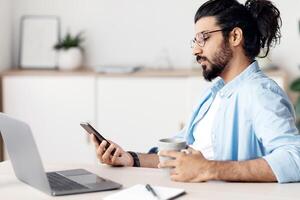Young Arab Male Freelancer Using Smartphone And Drinking Coffee, Relaxing At Workplace In Home Office, Sitting At Desk With Laptop Computer And Notepad, Resting After Finishing Project, Free Space photo