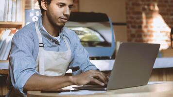 Work with accounts with social networks, online orders and payment of bills. Millennial african american male bartender in apron typing on laptop in modern cafe, cropped, panorama, free space photo