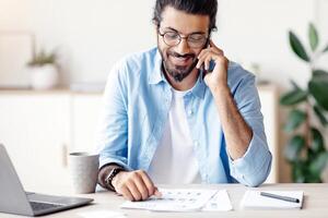 Busy Indian Freelancer Guy Checking Papers And Talking On Cellphone While Working On Laptop At Home Office, Speaking With Client, Sitting At Desk And Smiling, Having Plesant Phone Call, Free Space photo