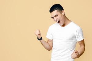 Happiness, expression of success, victory and good luck in business. Excited guy in white t-shirt shouts with joy of win and has fun, isolated on beige background, studio shot, free space photo