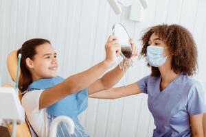 Good procedure and snow-white smile. Happy small girl looks in mirror for estimating dentist work and enjoying result with young african american specialist in uniform, protective mask and gloves photo