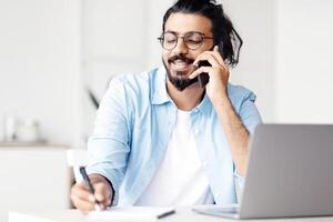 Young Arab Male Entrepreneur Talking On Cellphone And Taking Notes In Office, Handsome Eastern Manager Guy Sitting At Desk With Laptop And Noting Information To Notepad, Speaking With Client photo