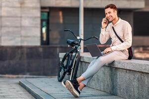 Busy guy solves business problems outside. Smiling man speaks at smartphone and works on laptop in city with bike photo