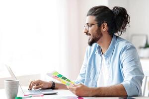 Handsome young eastern man interior designer holding color swatch samples and working on laptop at home office, checking palette for new design project, sitting at desk near window, copy space photo