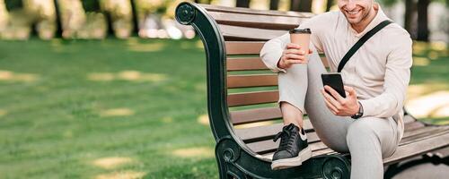 A young man sits on a park bench, sipping a coffee and looking at his smartphone. He appears relaxed, surrounded by greenery in a lively urban environment during daylight hours, copy space photo