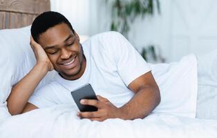 Smiling African Guy Using Cellphone Browsing Internet And Texting On Phone Lying In Bed At Home. Black Man With Smartphone Networking In Social Media. New Mobile App Advert Concept photo