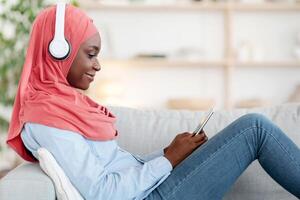 Technologies For Leisure. Black muslim lady in wireless headphones relaxing with smartphone at home, listening music and browsing internet, resting on comfortable couch in living room, side view photo