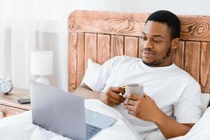 Happy Black Man Using Laptop Watching Movie Online And Having Coffee Enjoying Weekend Morning Lying In Bed At Home. Computer User Browsing Internet Searching Entertaining Web Content photo