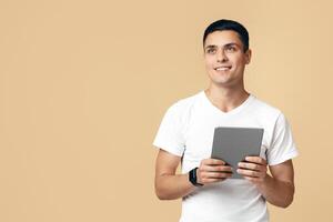 Management of modern technology, digital gadgets and social networks. Friendly young student blogger man with smart watch uses tablet and looks at empty space, isolated on light sand background photo