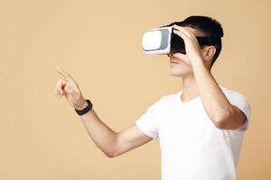 Man in goggles playing in games. Young student in white t-shirt and virtual reality spectacle touches to blank space with finger, isolated on sand or light background, side view, studio shot photo
