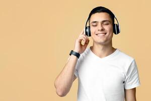 Enjoy music, stay at home. Smiling young blogger student man in white t-shirt with smart watch listens to song in modern headphones, isolated on light background photo