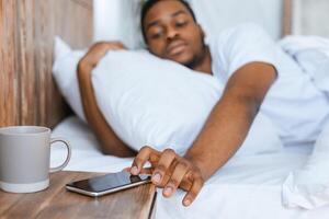 Sleepy Black Man Reaching For Smartphone Turning Off Alarm Clock Waking Up Early Lying In Bed In Bedroom At Home In The Morning. Lack Of Sleep And Rest Concept. Selective Focus, Shallow Depth photo