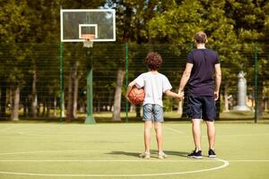 Healthy Lifestyle. Rear view of dad and son standing on playground with basket ball, holding hands, looking at hoop photo