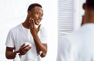 Handsome Black Man Using Facial Cream Moisturizing And Caring For Face Skin Standing Near Mirror In Bathroom Indoors In The Morning. Male Hygiene, Seflcare And Skincare Concept. Selective Focus photo
