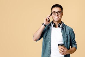 Coffee break, modern work and communication. Busy smiling young student man in glasses with headphones hold paper cup with coffee and talk on phone, isolated on light background, empty space photo