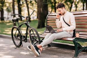 Important talk with client. Handsome young man with stubble in casual clothes working on laptop and speaks by phone while sitting on bench near bicycle in city, outdoors, side view, free space photo
