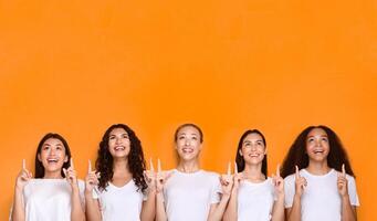 Five Diverse Girls Pointing Fingers Up Posing On Yellow Studio Background. Look There Concept. Copy Space photo