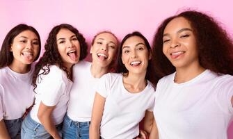 Five Multicultural Ladies Making Group Selfie Portrait Having Fun And Showing Tongues Posing Standing Over Pink Background, Smiling To Camera. Female Friendship Concept. Studio Shot photo
