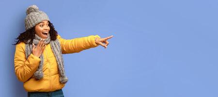Look There. Excited Black Woman In Winter Jacket Pointing Finger Aside At Copy Space Posing On Purple Studio Background. Panorama photo