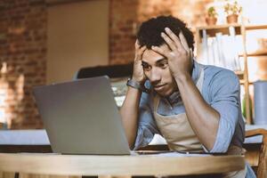 Ruin and financial problems during covid-19 quarantine. Sad young african american guy in apron holds head and looks in laptop at financial accounts and big debt in modern cafe, empty space photo