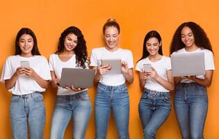 Five Cheerful Diverse Females Using Phones And Computers Networking Online And Browsing Internet Standing On Yellow Background, Studio Shot. Happy Ladies Posing With Different Gadgets photo