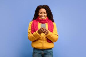 African American Woman Using Smartphone Phone Networking Texting In Social Media Mobile App Standing Over Purple Studio Background, Wearing Winter Clothes. Cellphone Application Concept photo