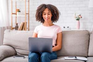 Online work in social networks during quarantine and self-isolation. Friendly african american girl working in laptop, sitting on couch with notepad photo