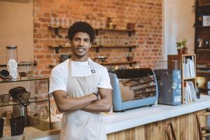 Opening of modern cafe after covid-19 lockdown. Young handsome african american guy in apron with crossed arms looking at camera in interior of modern loft cafeteria with equipment, empty space photo