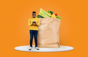 A customer stands with a paper grocery bag full of fresh produce while using a smartphone. Bright orange background highlights the new normal of shopping and delivery convenience. photo