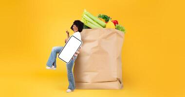 A customer happily engages in grocery shopping, holding a mobile phone in one hand while carrying a large paper bag filled with fresh produce. photo