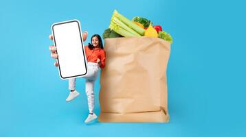 A happy customer is enjoying grocery shopping while holding a smartphone. The paper bag is filled with fresh vegetables and fruits, symbolizing modern shopping trends. photo