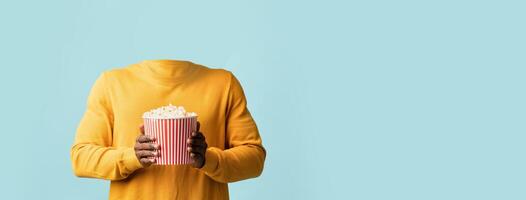 A headless figure dressed in a yellow sweater holds a large striped popcorn bucket against a soft blue background. This isolated composition emphasizes lifestyle themes and fun activities. photo