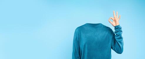 A headless individual dressed in a blue sweater makes a gesture of approval against a plain blue backdrop. This unique representation emphasizes a minimalist and surreal lifestyle concept. photo