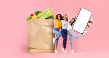Three friends joyfully pose in front of a large paper grocery bag filled with fresh produce, depicting a new normal in shopping. They hold a phone, reflecting remote grocery delivery trends. photo