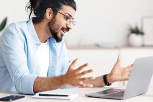Young Arab Freelancer Guy Making Call With Laptop At Home, Having Web Conference With Clients. Handsome Easter Guy Talking At Computer Camera, Enjoying Online Communication And Remote Work photo