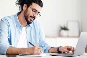 Young Indian Businessman Taking Notes While Working On Laptop Computer In Office, Western Freelancer Guy Noting Down Information From Internet, Planning Working Schedule, Sitting At Desk, Copy Space photo