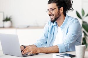 Smiling western freelancer guy working on laptop at home office, sitting at desk using computer, young indian man in eyeglasses typing on keyboard, looking at screen, enjoying remote work, copy space photo