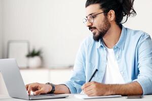 Young indian man entrepreneur using laptop and taking notes while working in modern office, sitting at desk and looking at computer screen, wester freelancer guy writing in notepad, copy space photo