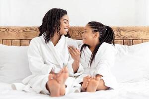 Joyful Black Mom And Daughter In Bathrobes Relaxing With Smartphone In Bed, Browsing Internet, Ordering Food Or Using Cellphone For Online Shopping, Cheerful Mother And Little Girl Having Fun Together photo