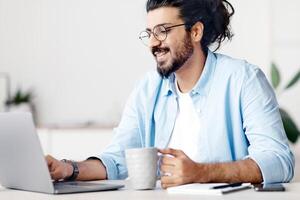 Pros of Freelancing. Positive Arab Freelancer Guy Drinking Coffee And Working On Laptop At Home Office, Eastern Man Sitting At Desk And Typing On Computer Keyboard, Enjoying Remote Job, Free Space photo
