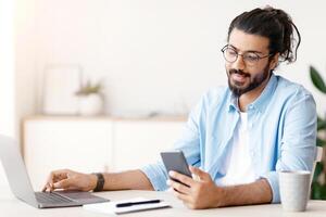 Millennial Arab Freelancer Guy Using Smartphone While Working On Laptop At Desk In Home Office, Browsing New Application Or Messaging With Friends, Enjoying Remote Job Opportunities, Copy Space photo