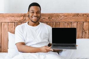 Happy African American Guy Showing Laptop With Empty Screen Advertising Internet Website Sitting In Bed At Home, Smiling To Camera. Computer User Advertising Internet Service Or Web Content. Mockup photo
