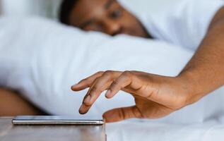Black Man Turning Off Alarm Clock On Phone Waking Up In The Morning Lying In Bed In Bedroom At Home. Focus On Cellphone. Early Awakening Concept. Shallow Depth photo