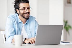 Young Indian Male Writer Using Laptop Computer, Sitting At Desk In Home Office, Handsome Western Man In Eyeglasses Typing On Keyboard And Looking At Screen, Enjoying Working Remotely, Selective Focus photo
