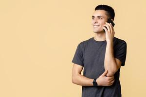 Call to friend or conversation about work, stay at home during COVID-19. Smiling young man student in brown t-shirt speaks on phone, looks at empty space, isolated on beige background, studio shot photo