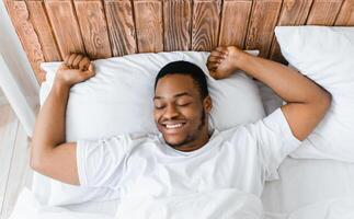 Rested African American Man Waking Up Stretching Hands Awakening Lying In Bed At Home In The Morning, Smiling With Eyes Closed. Good Sleep, Rest And Recreation Concept. Above View photo