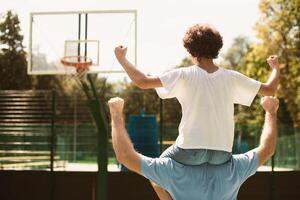 Celebrating Goal. Rear back view of adult father riding his son on shoulder, shaking clenched fists and screaming yes, making winner gesture, looking at basket, standing on court outdoors, cheering photo