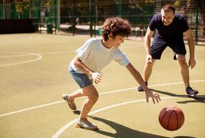 Men's Leisure. Curly child playing basketball on a sports field with his mature coach, copy space, blurred background photo